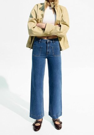 Femme portant une veste courte beige, un crop top blanc, un jean bleu taille haute à jambes larges, et des chaussures Mary Jane marron foncé cloutées.