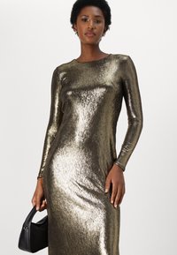 Metalická zlatá dlouhá šaty s upnutou siluetou, hladkou texturou a kulatým výstřihem, doplněné černou kabelkou.