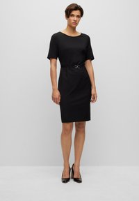 Black fitted dress with a round neckline, short sleeves, and a slim belt featuring a silver buckle. Smooth texture, knee-length design.