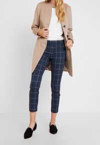 Beige langt frakke over en hvid top, parret med mørkeblå ternede bukser og sorte loafers. Bukserne har en slim fit og er ankellange.