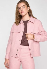 Veste en laine rose clair avec fermeture à boutons et deux poches poitrine. Texture lisse et coupe décontractée, associée à un haut marron et un pantalon assorti.