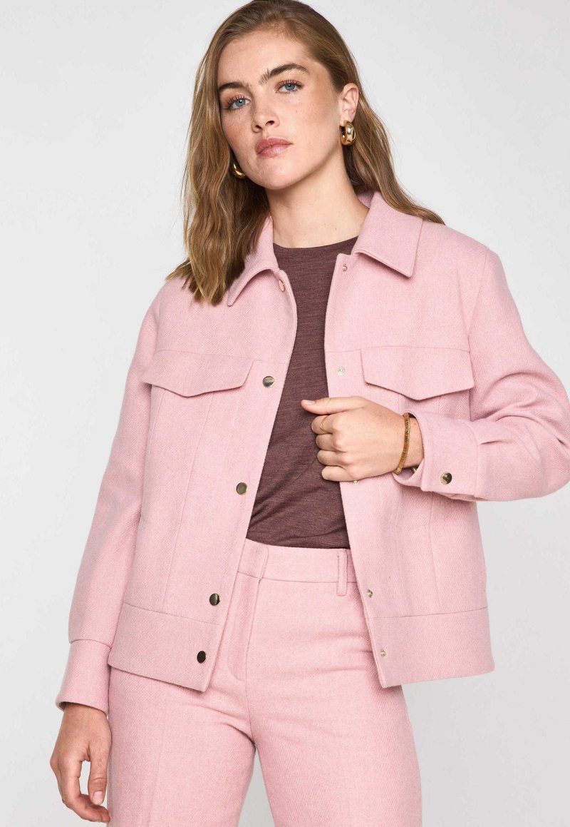 Veste en laine rose clair avec fermeture à boutons et deux poches poitrine. Texture lisse et coupe décontractée, associée à un haut marron et un pantalon assorti.