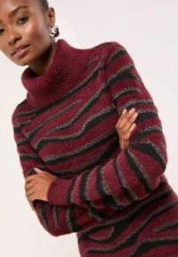 Bordowy sweter z golfem o miękkiej, puszystej fakturze. Posiada czarne i srebrne paski w faliste wzory, długie rękawy i wysoki kołnierz.
