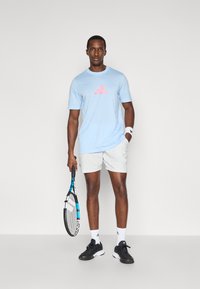 Jasnoniebieska koszulka sportowa z różowym graficznym logo, w zestawie z szarymi szortami. Model trzyma rakietę tenisową i ma na sobie czarne trampki.