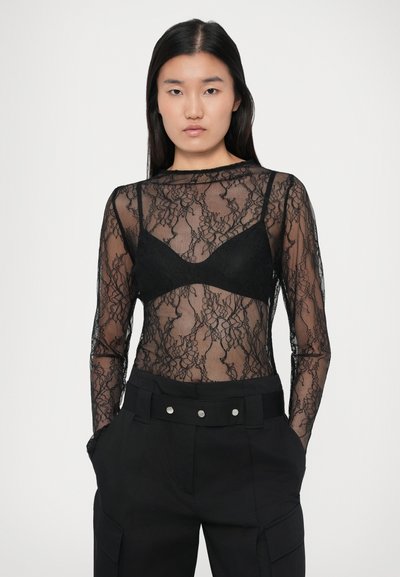 Femeie îmbrăcată într-o bluză din dantelă neagră transparentă cu mâneci lungi, având un bralette negru dedesubt și pantaloni negri cu talie înaltă și o curea lată.