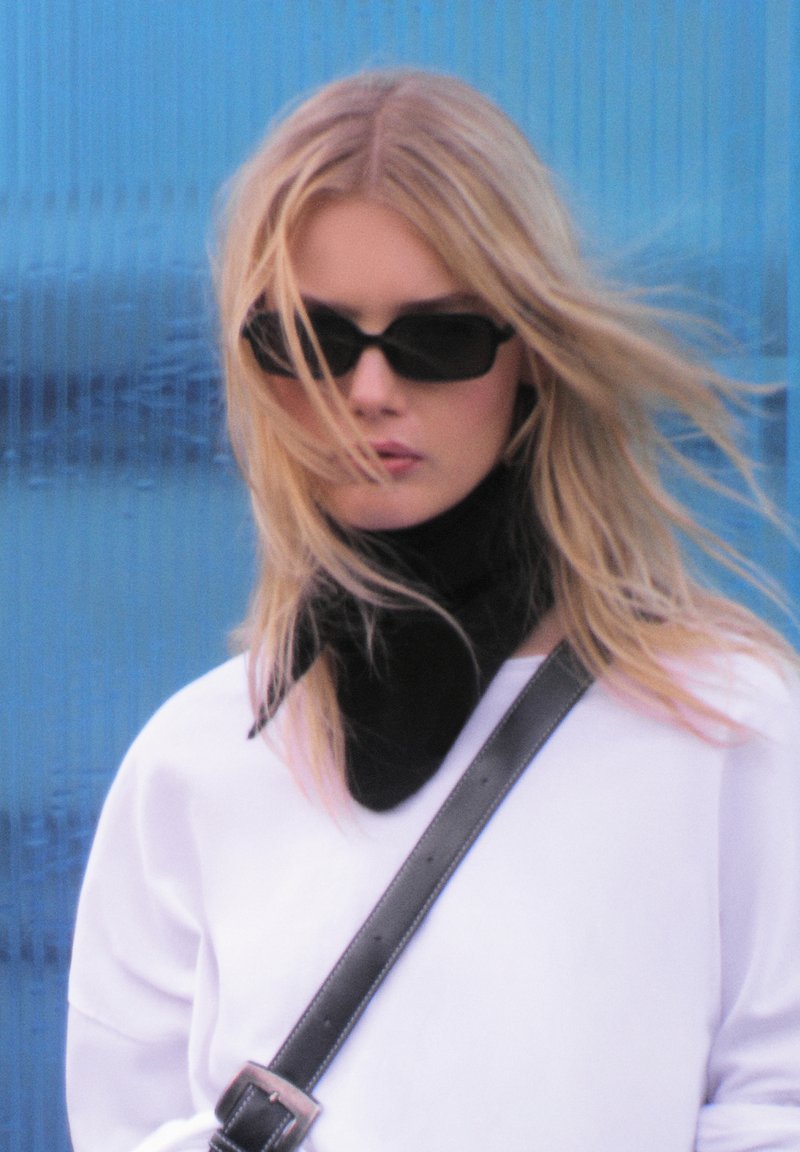 Black scarf, white oversized sweatshirt, and rectangular black sunglasses. Long, straight blonde hair flows in the wind against a blue background.