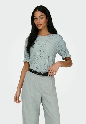 Kobieta o długich ciemnych włosach, ubrana w jasnoniebieską bluzkę z ażurowym wzorem i bufiastymi rękawami oraz szare spodnie z wysokim stanem i czarnym paskiem.