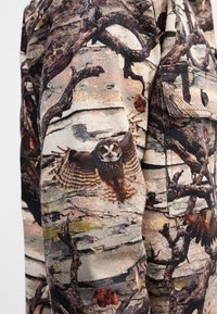 Camouflage jacket featuring a fabric with owl and branch patterns in earthy tones of beige, brown, and gray, with textured detailing.
