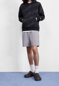 Schwarzer Hoodie mit weißem handschriftlichem Text, graue Shorts, weiße Socken und schwarze Clogs auf einem blauen Boden vor einem schlichten weißen Hintergrund.