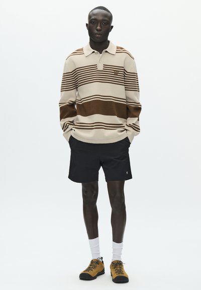 Polo à rayures beige avec des lignes horizontales marron, associé à un short noir, des chaussettes blanches et des chaussures de randonnée marron, posé sur un fond blanc.