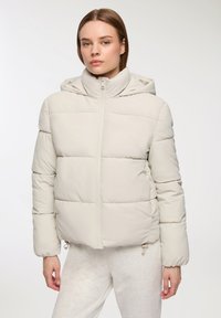 Jasno beżowa kurtka puchowa z zamkiem błyskawicznym i kapturem, z przeszywanymi sekcjami oraz regulowanymi sznureczkami na dole. Miękka, pikowana tekstura.