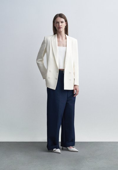 Femme portant un blazer blanc croisé, un haut blanc, un pantalon large bleu marine et des escarpins blancs à bout pointu, debout devant un fond clair uni.