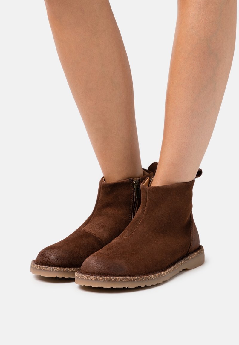 Paire de bottines en daim marron avec fermeture éclair latérale et semelles en caoutchouc, portées sur des jambes nues sur un fond clair uni.