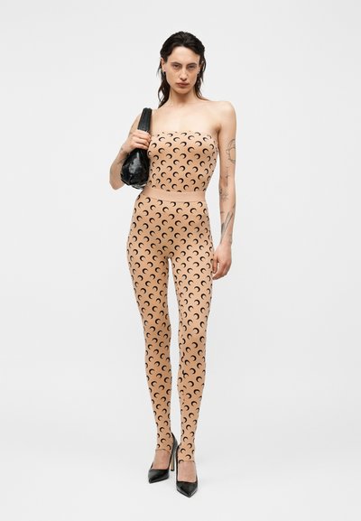 Ženska v bež brezrokavnem kombinezonu s črnim vzorcem polmeseca, črni visoki petki, drži črno torbico na ramenu, nevtralno ozadje.