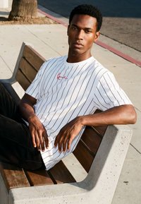 Ung mand med kort krøllet hår, der sidder på en træbænk udendørs, iført en hvid t-shirt med striber og sorte bukser.