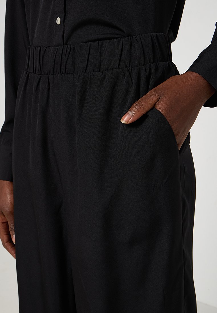 Zwarte elastische taillebandbroeken met zijzakken en een gladde stofstructuur, met een semi-loose pasvorm voor comfort en bewegingsvrijheid.