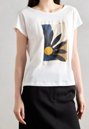 Wit shirt met korte mouwen en een abstracte bloemenprint in blauw, zwart, geel en beige, gedragen met een zwarte rok.