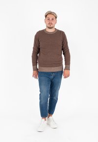 Maglione marrone lavorato a maglia, colletto rotondo, finitura testurizzata, orlo e polsini a coste, abbinato a jeans denim blu e sneakers bianche. Sfondo pulito.