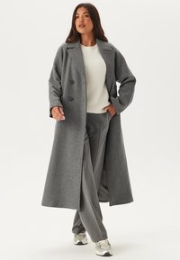 Cappotto grigio doppiopetto in lana, con grandi bottoni, maniche lunghe e un design a lunghezza media, abbinato a pantaloni grigi e sneakers.