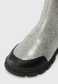 Šedé trblietavé členkové čižmy s čiernym gumovým špičkovým krytom a podrážkou. Značné textúrovaný povrch a kontúrovaný, vyvýšený tvar.