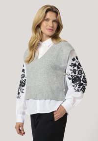 Gray knit sleeveless sweater layered over a white shirt with black floral embroidery on the sleeves, featuring ribbed texture and button accents.