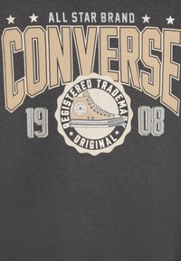 T-shirt nera con grande testo beige "CONVERSE", un logo circolare con un'immagine di sneaker e i numeri "19" e "08."