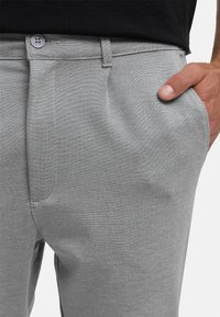 Mies, jolla on vaaleanharmaat housut, käsi osittain oikeassa taskussa, nappikiinnitteinen vyötärö ja musta paita housujen sisällä.