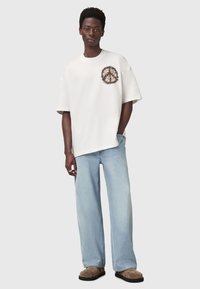 Hvid oversized t-shirt med et farverigt abstrakt design på brystet, parret med lyseblå denimbukser og brune slip-on sko.