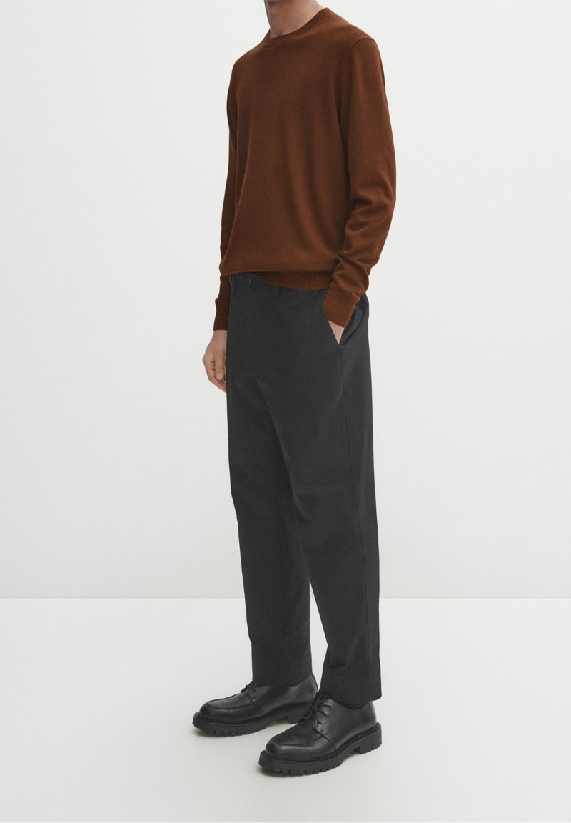 Bruine gebreide trui, relaxte pasvorm, ronde hals; zwarte op maat gemaakte broek, recht model; zwarte veterschoenen met dikke zolen.