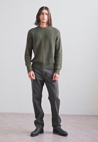 Dunkelgrüner strukturierter Pullover mit geripptem Ausschnitt, kombiniert mit grauen Jeans und schwarzen Stiefeletten, auf einem hellgrauen Hintergrund stehend.