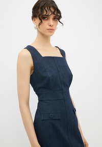 Jeanskleid in Dunkelblau mit quadratischem Ausschnitt und strukturiertem Oberteil. Mit seitlichen Taschen und Knopfdetails am Taillenbund.