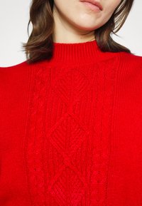 Czerwona sweter z wysokim dekoltem, teksturowanym wzorem w warkocze i prążkowanymi detalami na rękawach i dolnej części. Miękka dzianina.