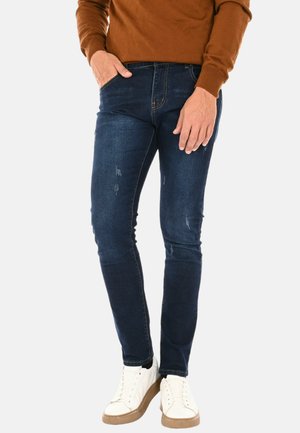 Jeans slim fit blu scuro con dettagli distressed, realizzati in denim, abbinati a sneakers bianche e un maglione marrone.