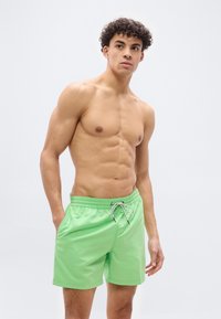 Νεαρός άνδρας με σγουρά μαλλιά, φορώντας ανοιχτό πράσινο μαγιό, στέκεται με το ένα χέρι στην τσέπη, κοιτάζοντας ελαφρώς προς το πλάι μπροστά σε απλό φόντο.