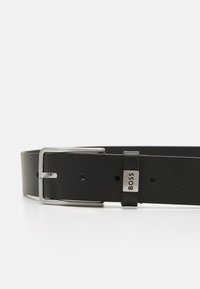 Ceinture en cuir noir avec une finition texturée, boucle en métal argenté et un petit accent métallique arborant le nom de la marque "BOSS."