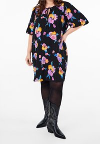 Femme portant une robe florale noire avec des fleurs violettes et oranges, des collants noirs et des bottes de cowboy noires à mi-mollet à clous, la main sur la hanche.