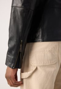 Μαύρο δερμάτινο μπουφάν με λεία υφή, διαθέτοντας πλευρικό φερμουάρ, συνδυασμένο με ανοιχτό μπεζ cargo παντελόνι που έχει μπροστινές τσέπες.