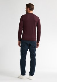 Burgunderfarbener langärmliger Pullover mit Rundhalsausschnitt, kombiniert mit dunkelblauen Hosen und weißen Sneakers, von hinten betrachtet.