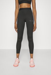 Leggings sportivi neri con un design aderente e lunghezza a metà polpaccio. Presentano un logo Puma e una cintura texturizzata. Indossati con scarpe sportive rosa.