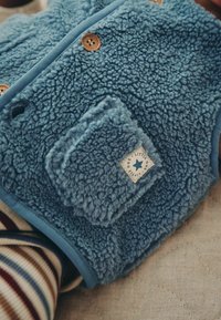 Blue fuzzy vest with wooden buttons and a small pocket labeled "Little One" worn over striped clothing.