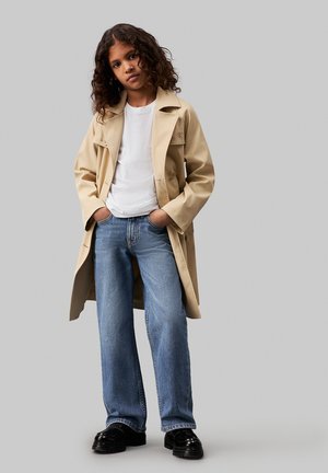 Trench coat beige con grande collare e bottoni, indossato sopra una maglietta bianca e jeans blu. Scarpe nere completano l'outfit, in piedi contro uno sfondo grigio.