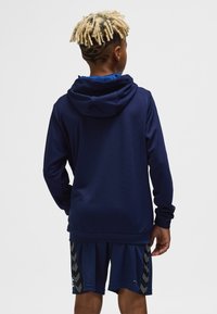 Marineblå hættetrøje med en struktureret overflade, der har hætte og lange ærmer, båret over matchende marineblå shorts med et chevronmønster på siderne.