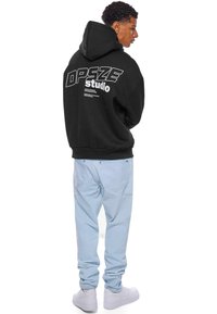 Sort hættejakke med hvid grafisk tekst på ryggen, kombineret med lyseblå jeans og hvide sneakers. Stoffet ser blødt og afslappet ud.