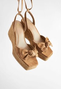 Braune Wildleder-Wedgeschuhe mit eckiger Zehenpartie und einem geknoteten Schleifendetail an der Vorderseite. Der Wedge-Absatz hat eine gewebte Textur. Verstellbare Knöchelriemen.