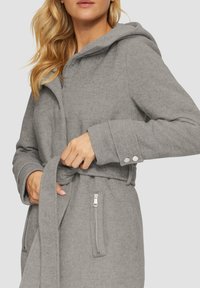Manteau à capuche gris en tissu texturé, ceinture à nouer à la taille, deux poches zippées et boutons-pression aux poignets.