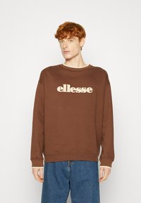 Sudadera marrón de cuello redondo con un logo "ellesse" en beige. Confeccionada en tela suave con puños y dobladillo acanalados. Combinada con unos jeans de mezclilla azules.