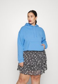 Sweatshirt azul com capuz e bolso canguru, combinado com uma saia preta em camadas com estampado floral, apresentando acentos de flores brancas sobre um fundo texturizado.