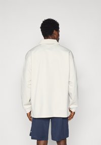 Polo branco de manga longa com colarinho, apresentando um corte descontraído e ombros caídos, combinado com calções azul-marinho. Textura suave, design minimalista.