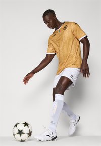Dres s futbalom s textúrou zlata a bielymi akcentmi, doplnený o logo, v kombinácii s bielymi šortkami a ponožkami po kolená, pričom noha má pätu s futbolom.