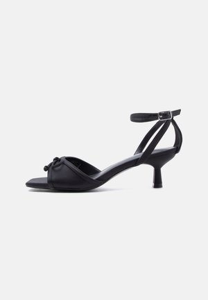 Black open-toe heeled sandal with ankle strap, small bow detail on toe strap, and a low tapered heel against a plain background.